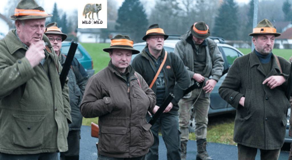 Gruppe von Hobby-Jägern in Jagdkleidung mit Waffen, im Hintergrund Fahrzeuge und Bäume.