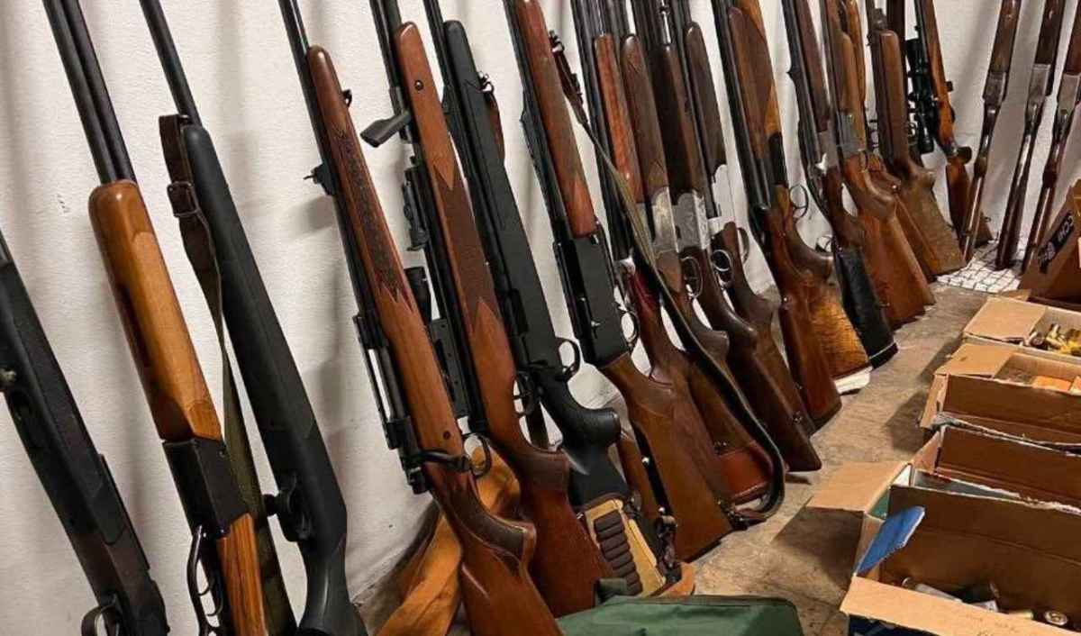 Decenas de escopetas y rifles de caza apoyados contra una pared blanca, junto a cajas de munición