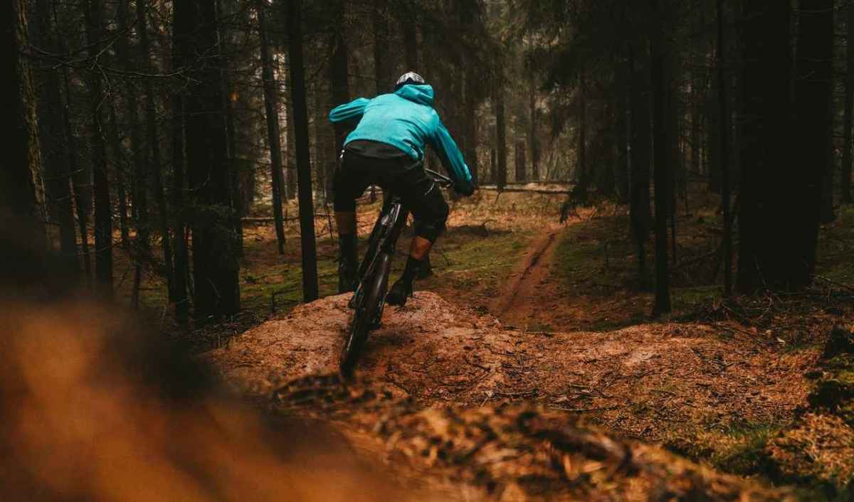 Mountainbiker mit türkisfarbener Jacke fährt auf einem Waldweg zwischen dunklen Nadelbäumen